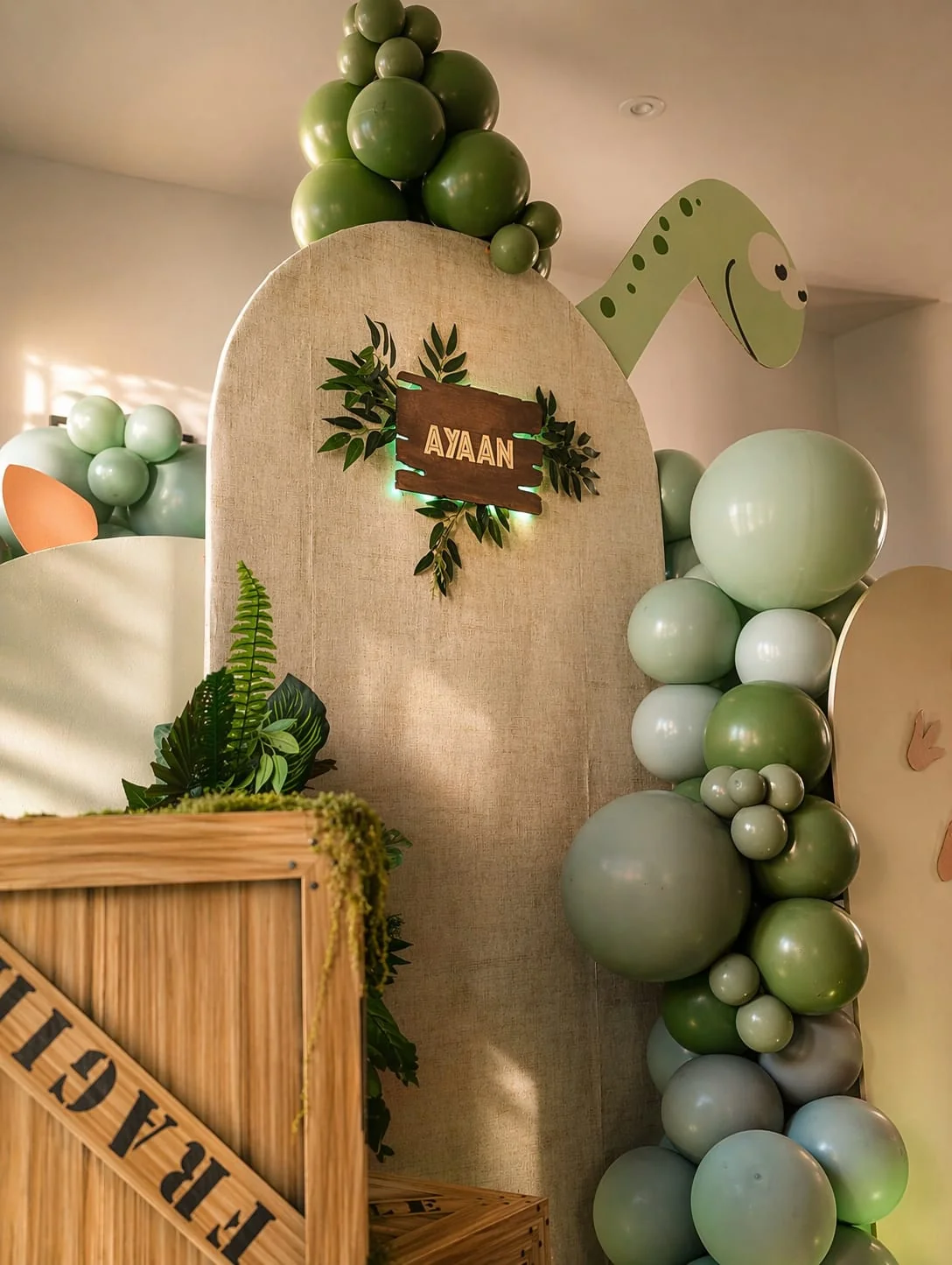 Close-up detail of an illuminated AYAAN wooden name sign nestled in greenery, surrounded by sage and green balloons, on a neutral arch panel
