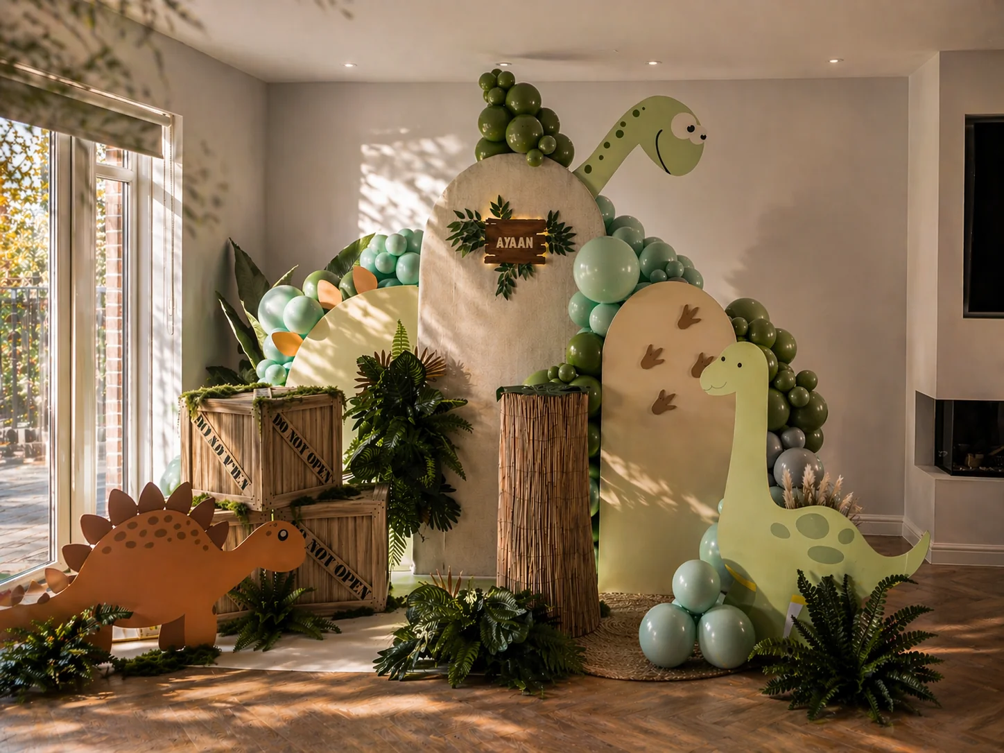 Finished dinosaur themed children's party setup in a Surrey home, with sage balloon arch, AYAAN name sign, cardboard brontosaurus and stegosaurus cutouts, wooden explorer crates and tropical foliage