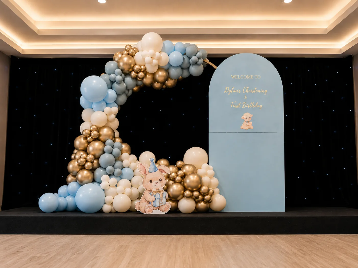 Sky blue, chrome gold and ivory balloon hoop beside an arched welcome backdrop against a black starlight wall