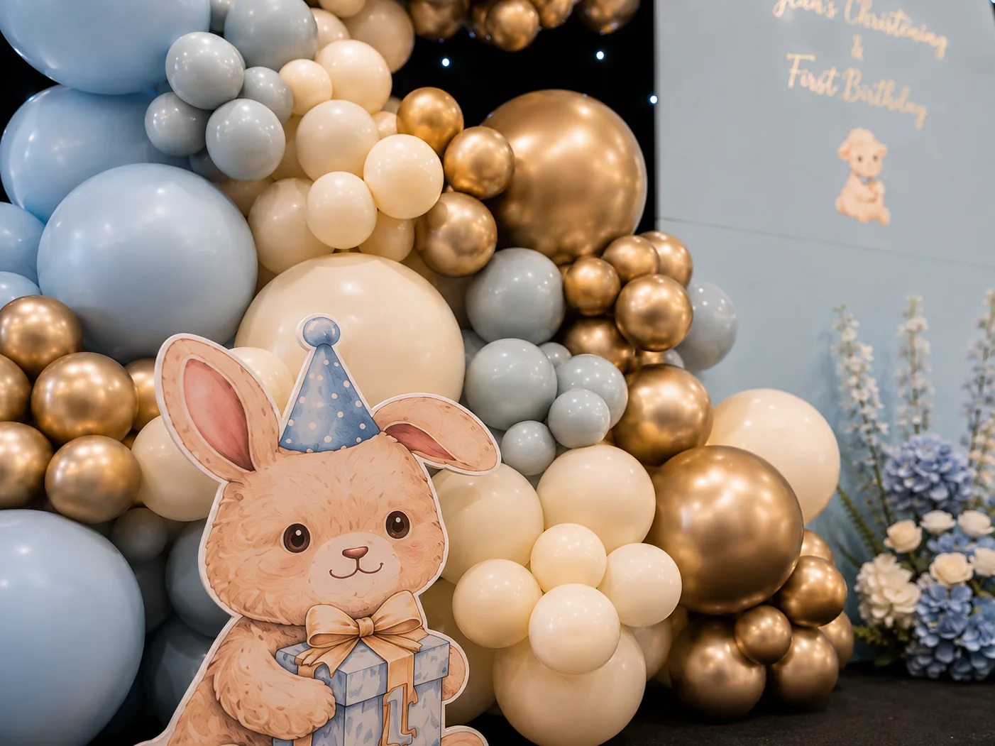 Painted rabbit cutout in a party hat holding a gift box at the foot of a chrome gold, sky blue and ivory balloon cluster