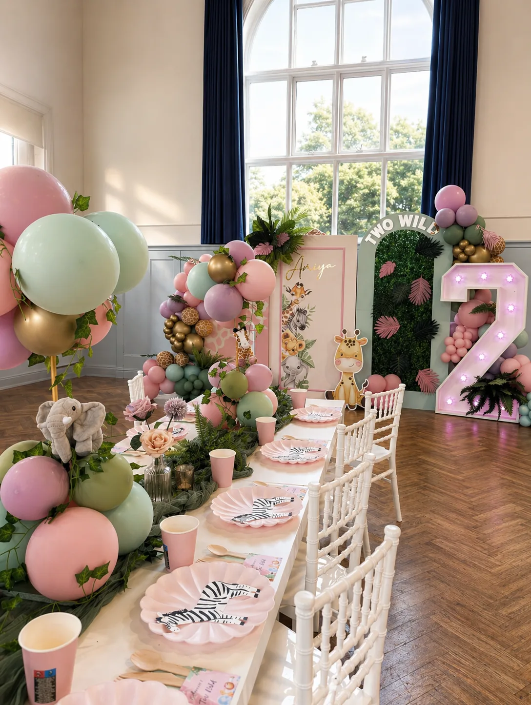 Finished Two Wild safari second birthday styling at Horley Community Centre in Surrey by River Fox Events — pastel balloon garland with foliage, personalised Amiya safari name panel, marquee 2 with pink bulb lights, twelve seat banquet table dressed with foliage runner, plush elephant centrepiece, pink scallop plates, zebra print salad plates and gold cutlery